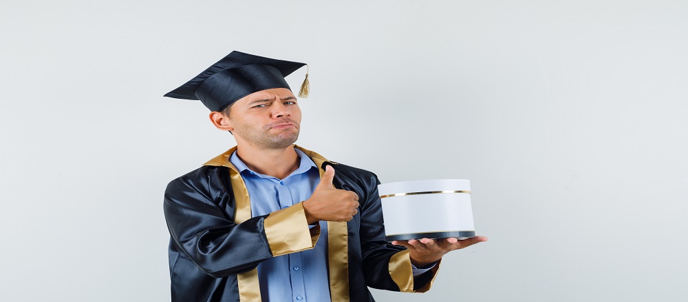 young male holding gift box while showing thumb up in graduation gown,cap and looking glad , front view.